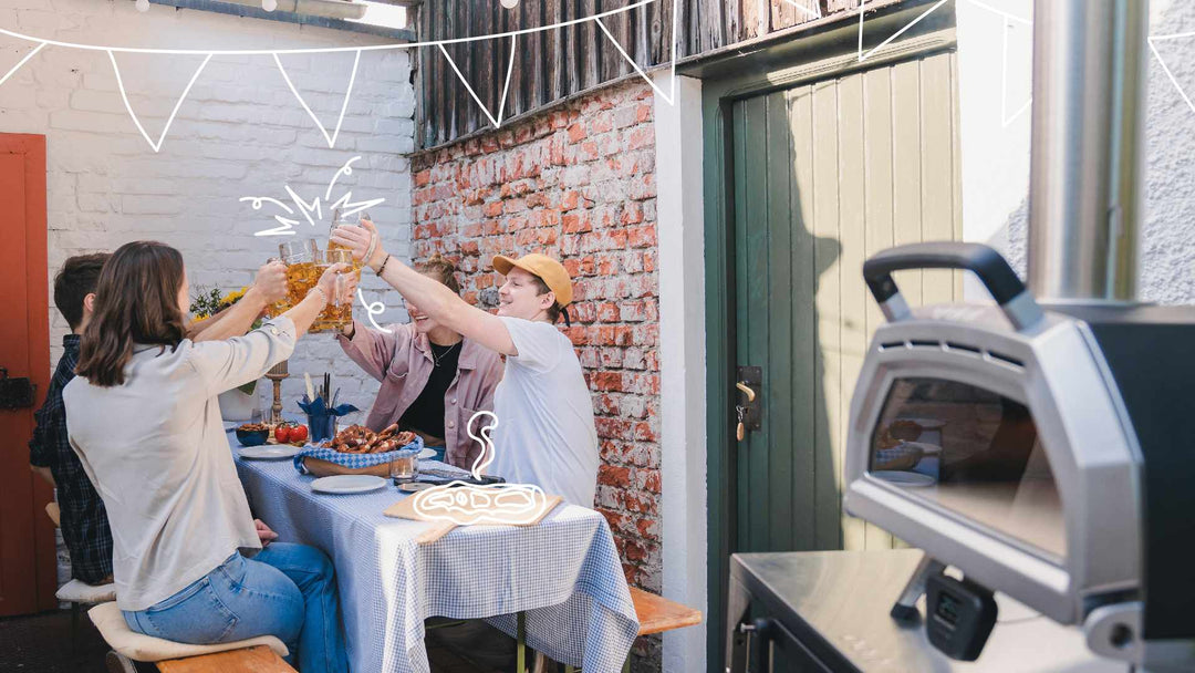 Four people sitting at a table outside and cheering with glasses in the background, a Ooni Karu 16 Multi-Fuel Pizza Oven in the foreground.