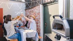 Four people sitting at a table outside and cheering with glasses in the background, a Ooni Karu 16 Multi-Fuel Pizza Oven in the foreground.
