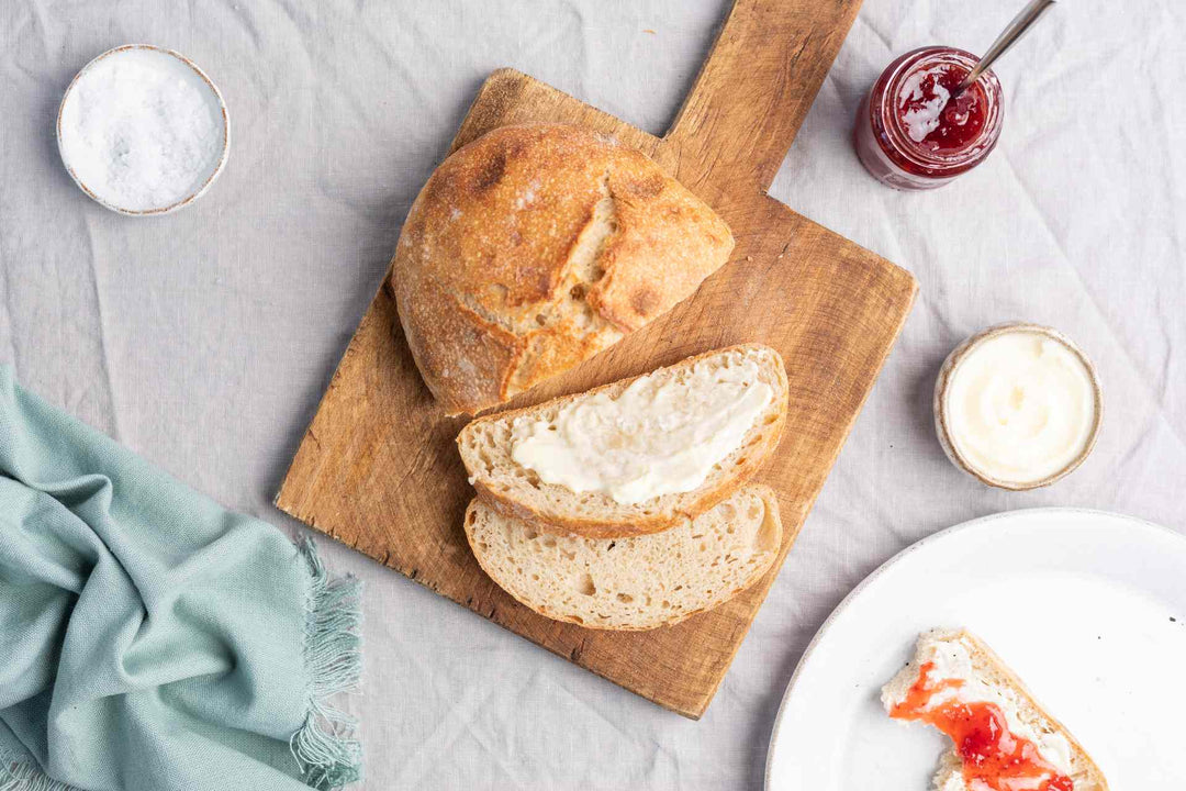 Half of a fresh-baked, no-knead bread loaf and two slices, one spread with butter, all on a bamboo serving board next to a plate 