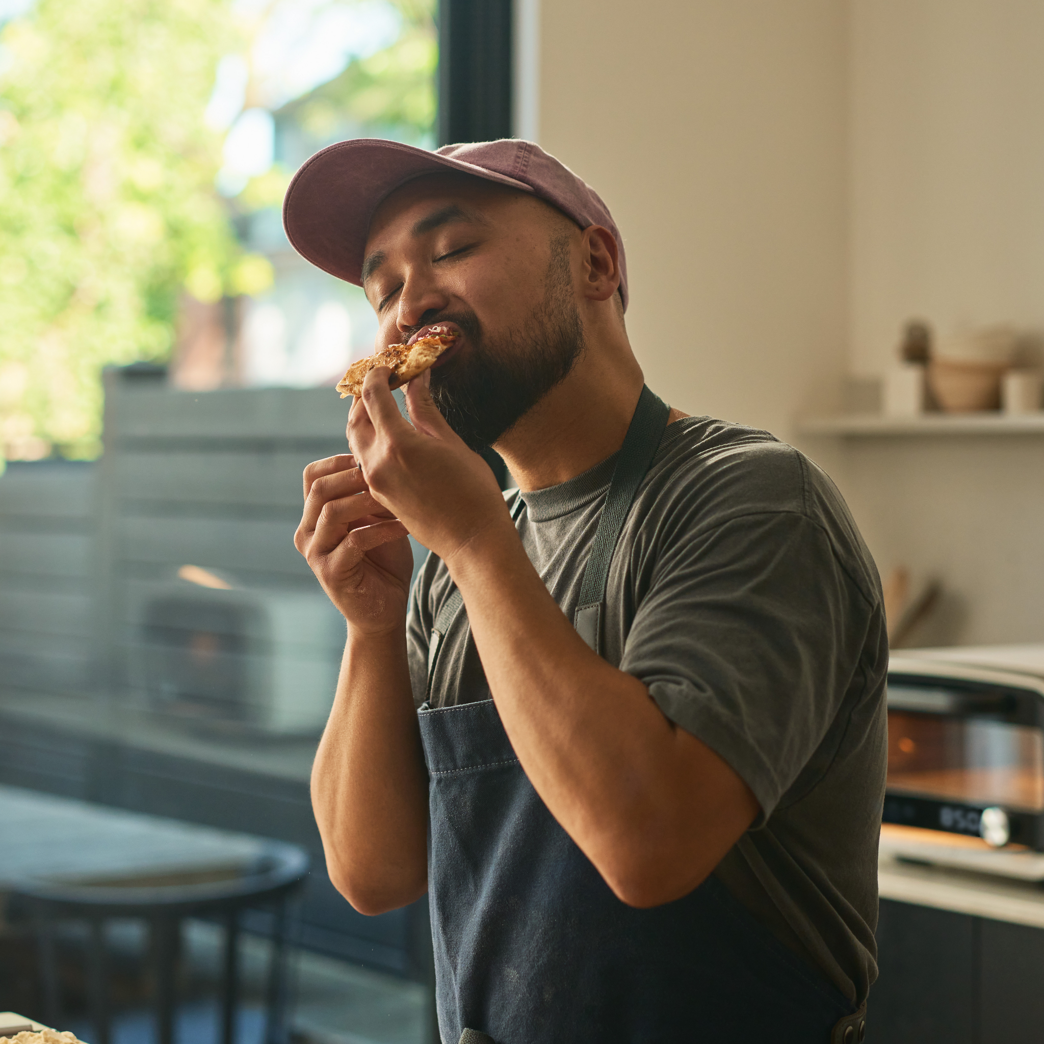 person eating a slice of pizza in kitchen. Ooni Volt in the background